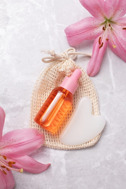 Bouteille de sérum sans marque sur un sac en coton avec des fleurs de lys sur fond de marbre Pierre Gua Sha pour la massothérapie faciale Traitement anti-âge et tonifiant