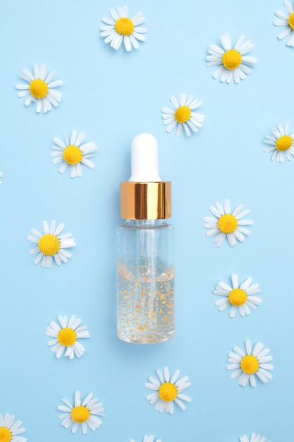 Bouteille transparente avec sérum de beauté acide hyaluronique or 24 carats et vitamines avec des fleurs de camomille sur fond bleu Concept de spa soins du visage et du corps