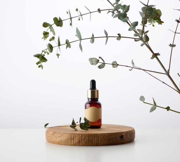 Une bouteille en verre marron avec une pipette sur un podium en bois et une branche d'eucalyptus fond blanc Publicité et promotion de produits cosmétiques