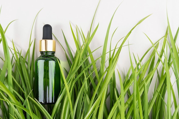 Une bouteille en verre vert avec sérum, huile essentielle, collagène ou autre produit cosmétique parmi l'herbe verte sur fond blanc. Maquette de concept cosmétique de Spa biologique naturel Vue de dessus.