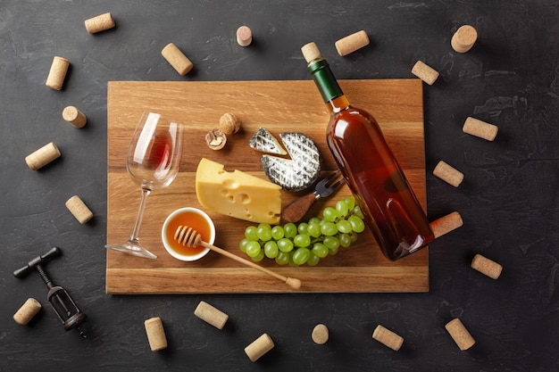 Bouteille de vin blanc, tête de fromage, grappe de raisin, miel, noix et verre à vin sur une planche à découper avec tire-bouchon