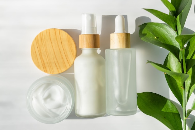 Bouteilles cosmétiques blanches et pot ouvert avec crème pour le visage sur fond blanc avec des feuilles de ruscus vert Concept de beauté pour le traitement du corps Soins de la peau Emballage sans marque pour le traitement de la peau