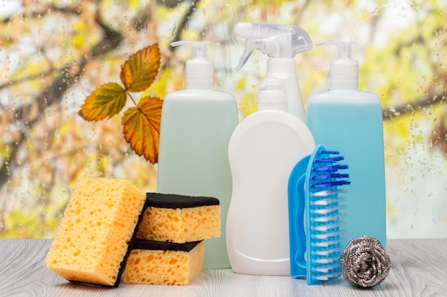 Des bouteilles de liquide vaisselle, des éponges et une brosse devant la fenêtre avec des gouttes d'eau et des feuilles d'automne. Concept de lavage et de nettoyage.