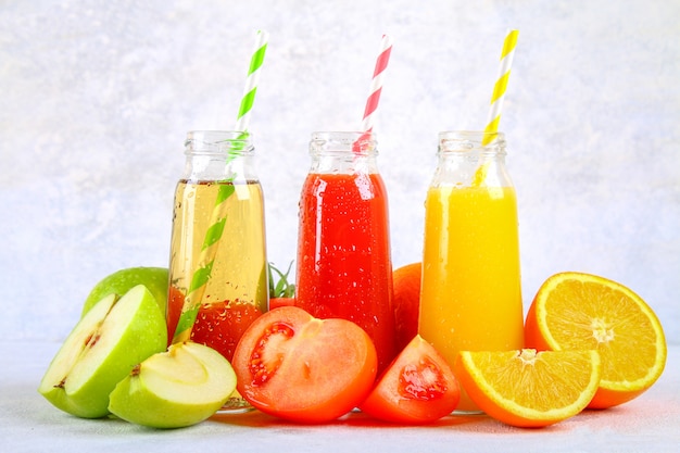 Bouteilles avec orange fraîche, pomme, jus de tomate et tubules colorés sur une table en béton grise.