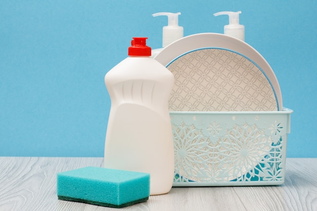 Bouteilles en plastique de liquide vaisselle, nettoyant pour vitres et carreaux, assiettes propres dans le panier et éponge sur fond bleu. Concept de lavage et de nettoyage.