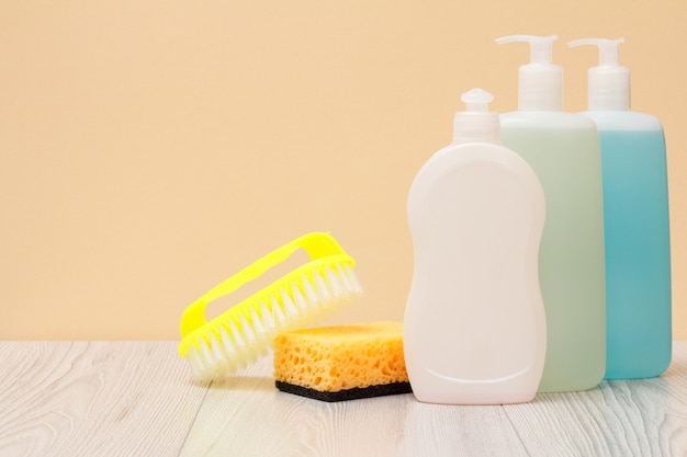 Bouteilles en plastique de liquide vaisselle, nettoyant pour vitres et carreaux, brosse et éponge sur planches de bois et fond beige. Concept de lavage et de nettoyage.