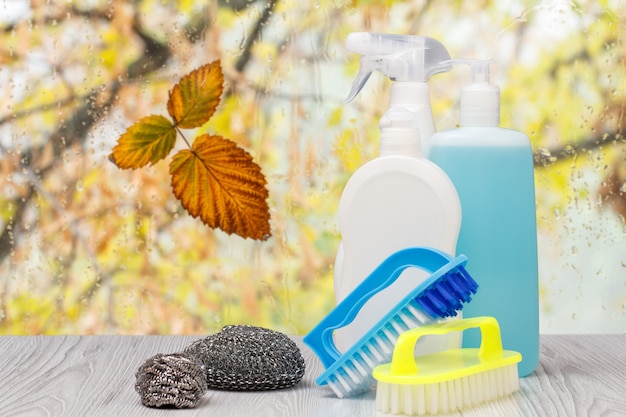 Bouteilles en plastique de liquide vaisselle, nettoyant pour vitres et carreaux, brosses et éponges métalliques devant la fenêtre avec des gouttes d'eau et des feuilles d'automne. Concept de lavage et de nettoyage.