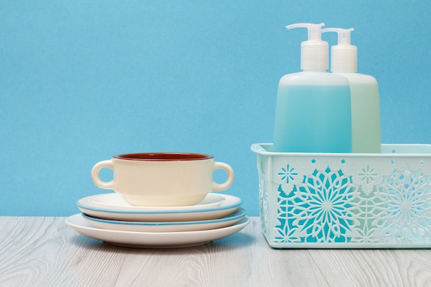 Bouteilles en plastique de liquide vaisselle, nettoyant pour vitres et carreaux dans le panier, assiettes propres et bol sur fond bleu. Concept de lavage et de nettoyage.