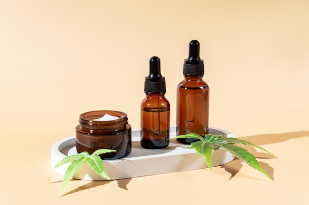 Bouteilles en verre avec huile de cbd à base de plantes et crème pour le visage ou teinture et feuille de chanvre sur fond beige pastel Huile de cbd cosmétique Huiles naturelles essentielles pour le visage et le corps