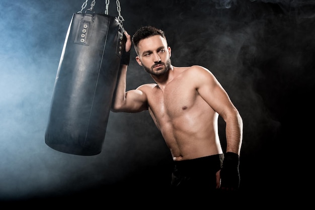 Boxeur athlétique sérieux tenant un sac de boxe sur fond noir avec de la fumée