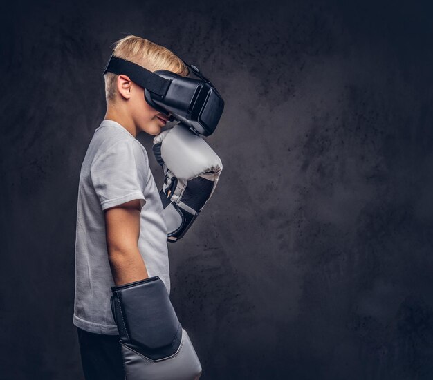 Boxeur écolier aux cheveux blonds vêtu d'un t-shirt blanc portant des lunettes de réalité visuelle et des gants de boxe, entraînement en studio. Isolé sur un fond texturé sombre.