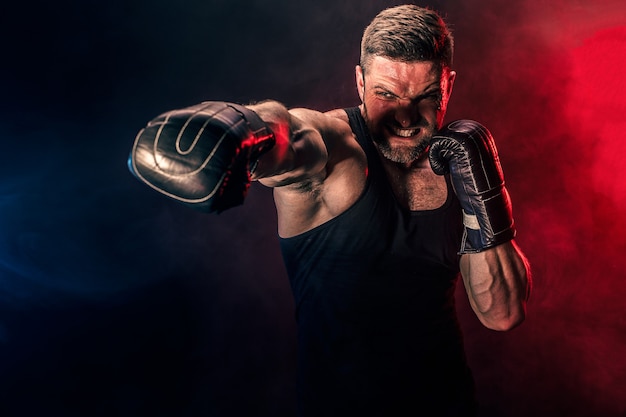 Boxeur muay thai sportif tatoué barbu en maillot de corps noir et gants de boxe combattant sur fond sombre avec de la fumée. Concept de sport.