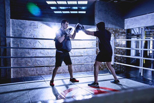 Photo des boxeurs professionnels avec des gants s'entraînent à des combats dans des couleurs sombres de ring de boxe en salle