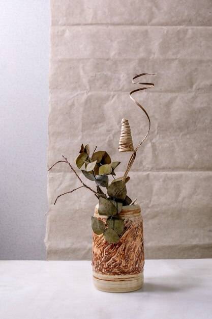 Branche de fleurs sèches, d'eucalyptus et de brindilles dans un vase en céramique marron sur une table en marbre blanc avec un mur gris derrière. C