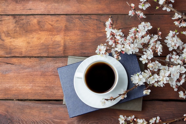 Branches de Sakura avec fleurs, tasse blanche avec café noir et livre sur une surface en bois sombre. Mise à plat, vue de dessus