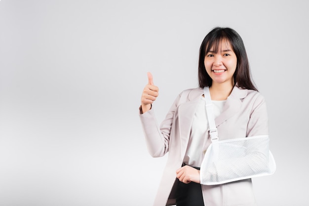 Bras cassé. Femme d'affaires sourire confiant cassé après un accident porter une attelle de bras pour le traitement montrer le doigt vers le haut pour un bon signe, main de soutien de fronde femme asiatique heureuse isolée sur fond blanc