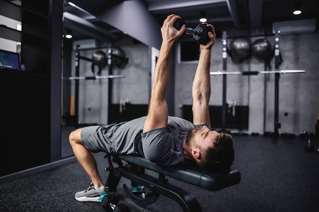 Bras et poitrine de pompage. Un jeune homme séduisant en chemise grise lève un haltère au-dessus de sa tête dans la salle de sport. Force, mouvement puissant. Posture initiale du corps et discipline sportive