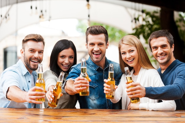 Bravo les amis ! Groupe de jeunes heureux se liant les uns aux autres et étirant des bouteilles avec de la bière tout en se tenant à l'extérieur