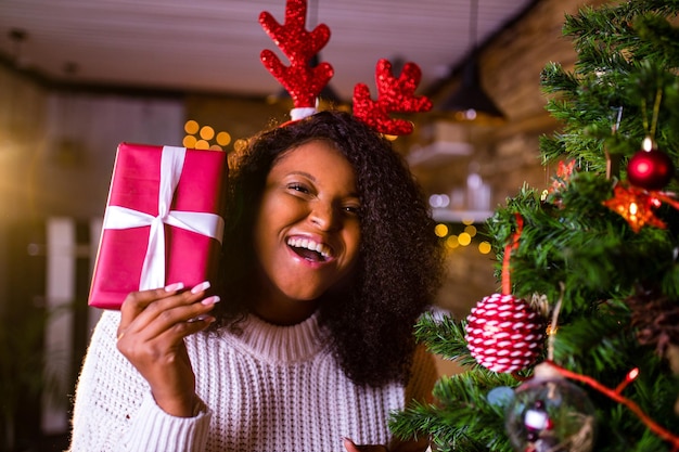 Brésil brunette hair woman hold red gift box près de firtree dans son appartement, elle porte un pull blanc et des cornes de cerf rouge