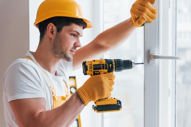 Bricoleur en uniforme jaune installe une nouvelle fenêtre à l'aide d'un tournevis automatique Conception de rénovation de maison
