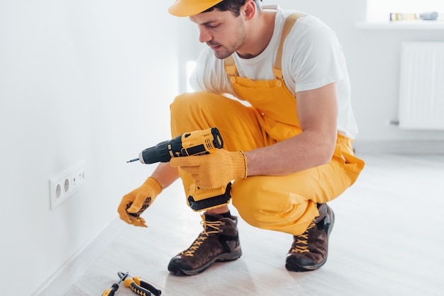 Bricoleur en uniforme jaune travaille avec l'électricité et installe une nouvelle prise à l'aide d'un tournevis automatique Conception de rénovation de maison