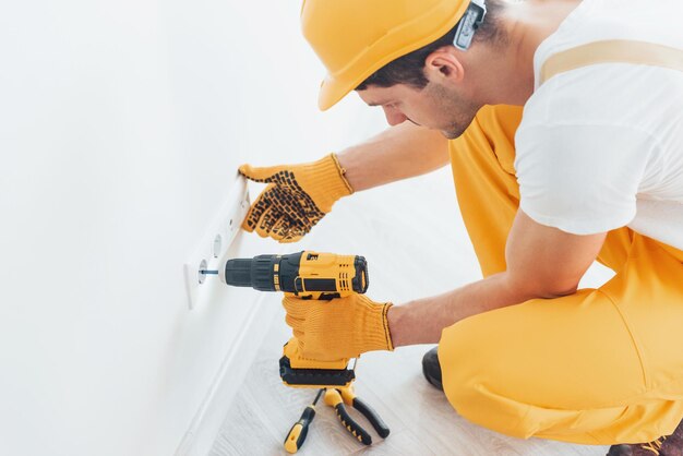 Bricoleur en uniforme jaune travaille avec l'électricité et installe une nouvelle prise à l'aide d'un tournevis automatique Conception de rénovation de maison