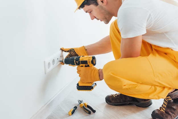 Bricoleur en uniforme jaune travaille avec l'électricité et installe une nouvelle prise à l'aide d'un tournevis automatique Conception de rénovation de maison