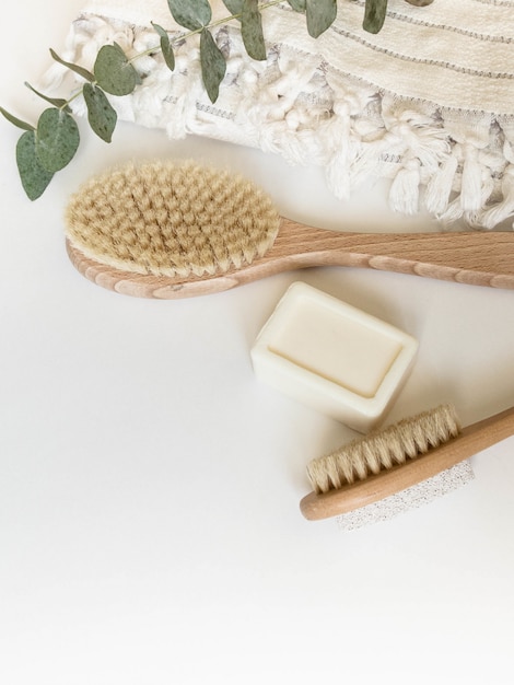 Brosse pour le corps avec manche en bois, ponce, serviette blanche et morceau de savon sur fond blanc. Vue de dessus.