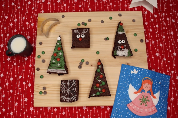 Photo des brownies en forme d'arbre de noël, du père noël, des rennes, une table en bois, des gouttes de chocolat et du lait.