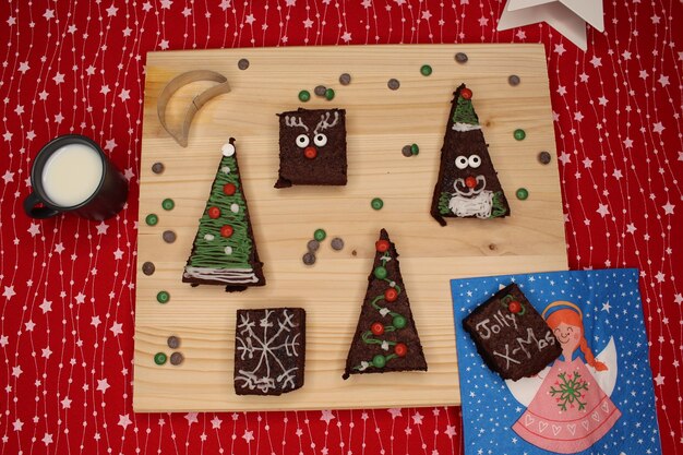 Photo des brownies en forme d'arbre de noël, du père noël, des rennes, une table en bois, des gouttes de chocolat et du lait.