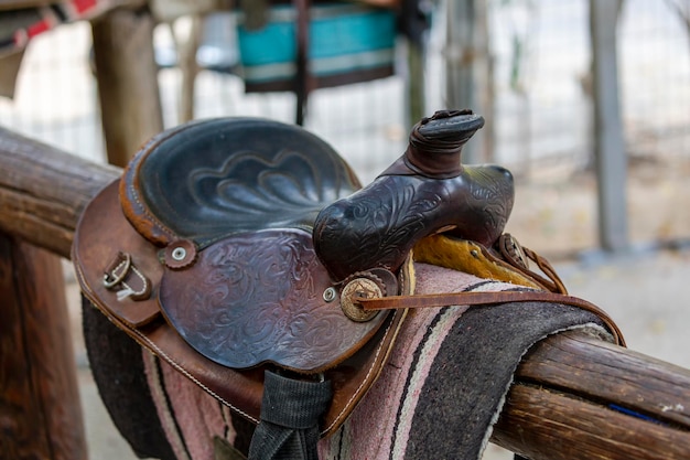 Brun de selle sur clôture contre le mur en bois. Selle de sport en cuir, harnais pour chevaux à la lumière du jour. Selles de cheval western sur un rack, prêtes pour l'entraînement de dressage. Fond de sport équestre, vintage