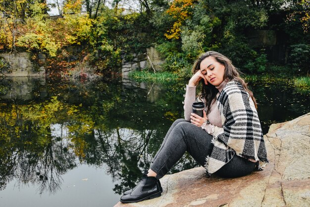 Brune aux cheveux longs assise au bord du lac et buvant du thé chaud dans un thermos. Repos d'automne en plein air. Nature d'automne.