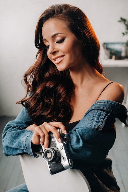 Une brune aux cheveux longs pose dans un studio assis sur une chaise avec un appareil photo dans ses mains.