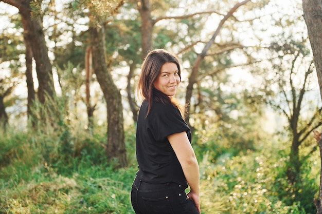 Brune en chemise noire et jeans debout et posant dans la forêt pendant la journée avec un beau soleil