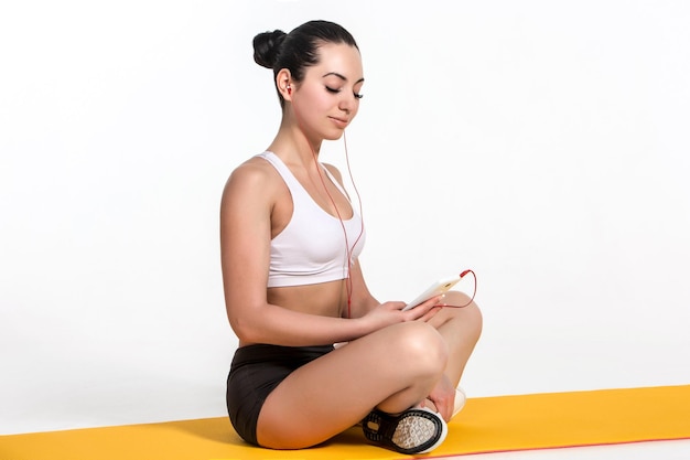 Brune avec un corps en forme sur un tapis de yoga. fille avec téléphone et casque. Mode de vie sain et concept sportif.