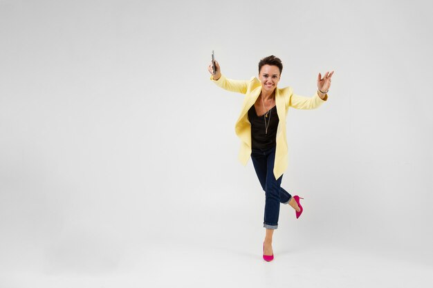 Photo brune avec une coupe courte dans une veste de couleur crème et des pompes roses pompes danse avec un téléphone dans ses mains sur gris