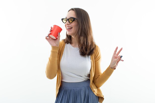 Brune dans un chandail jaune boit du café dans une tasse rouge Jeune femme en vêtements élégants sur fond blanc