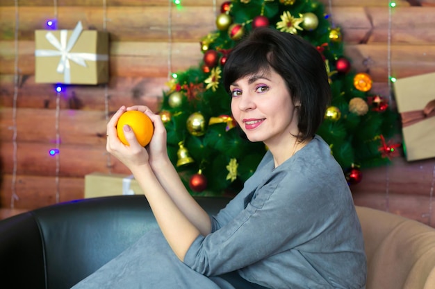 Une brune heureuse avec un sourire est assise sur le canapé dans une robe grise avec un orange vif dans les mains sur fond d'un bel arbre de Noël