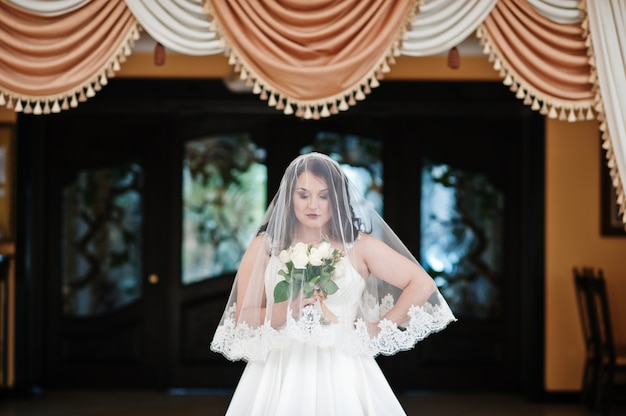 Brune mariée aux gros seins avec bouquet de mariée posé sous le voile à la salle de mariage