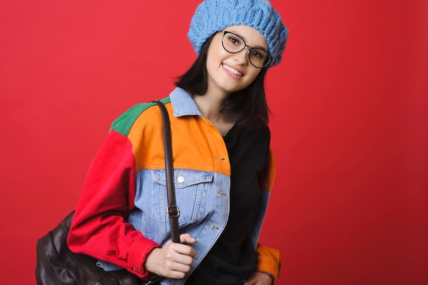 Brune moderne en veste en jean colorée et chapeau debout avec sac à dos sur rouge