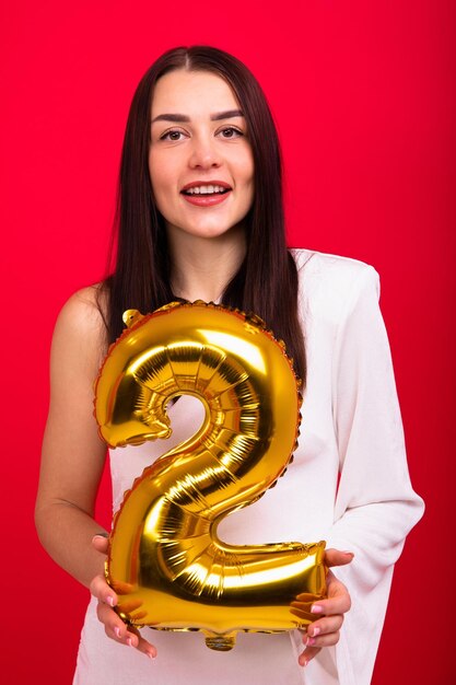 Une brune en robe blanche tient une boule en forme de numéro deux sur fond rouge Le concept de faire une année en chiffres