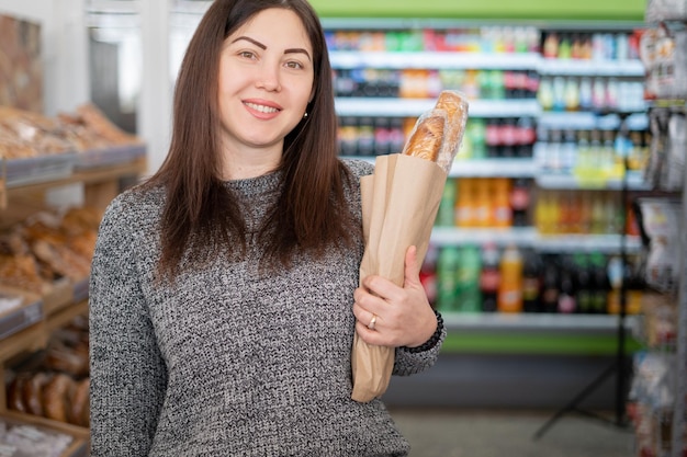 Une brune se tient sur les étagères du magasin en souriant et tenant du pain de blé dans un sac en papier