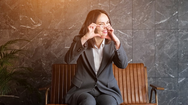 Une brune souriante en costume classique gris parle et gesticule assise sur un banc marron près d'une grande plante en pot lors d'un entretien d'embauche dans un nouveau bureau, lumière du soleil