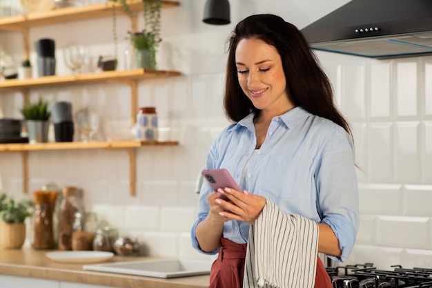 Une brune souriante est debout dans la cuisine et regarde quelque chose sur son smartphone une belle heure