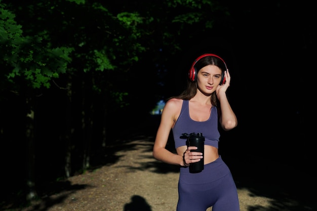 Une brune sportive avec des écouteurs et une bouteille d'eau après avoir fait du jogging dans le parc Entraînements fastidieux à l'extérieur