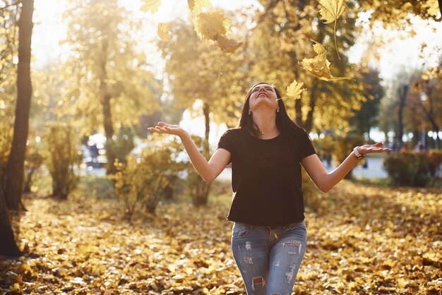 Une brune en tenue décontractée s'amuse avec des feuilles dans un magnifique parc d'automne.