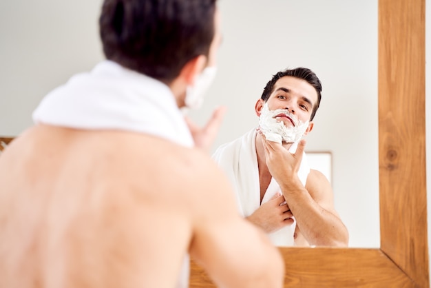 Brunet mâle avec de la mousse sur la barbe près du miroir le matin