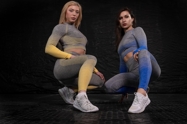 Brunette et blonde physique athlétique accroupie posant sur un fond sombre. vêtements de fitness. concept de mode de vie sain