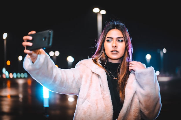 Brunette caucasienne prenant un selfie au téléphone, vêtue d'une veste en laine rose dans un parking vide. Session urbaine de nuit en ville