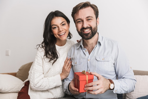 Brunette couple homme et femme tenant une boîte de cadeau d'anniversaire alors qu'il était assis sur un canapé dans une pièce lumineuse à la maison